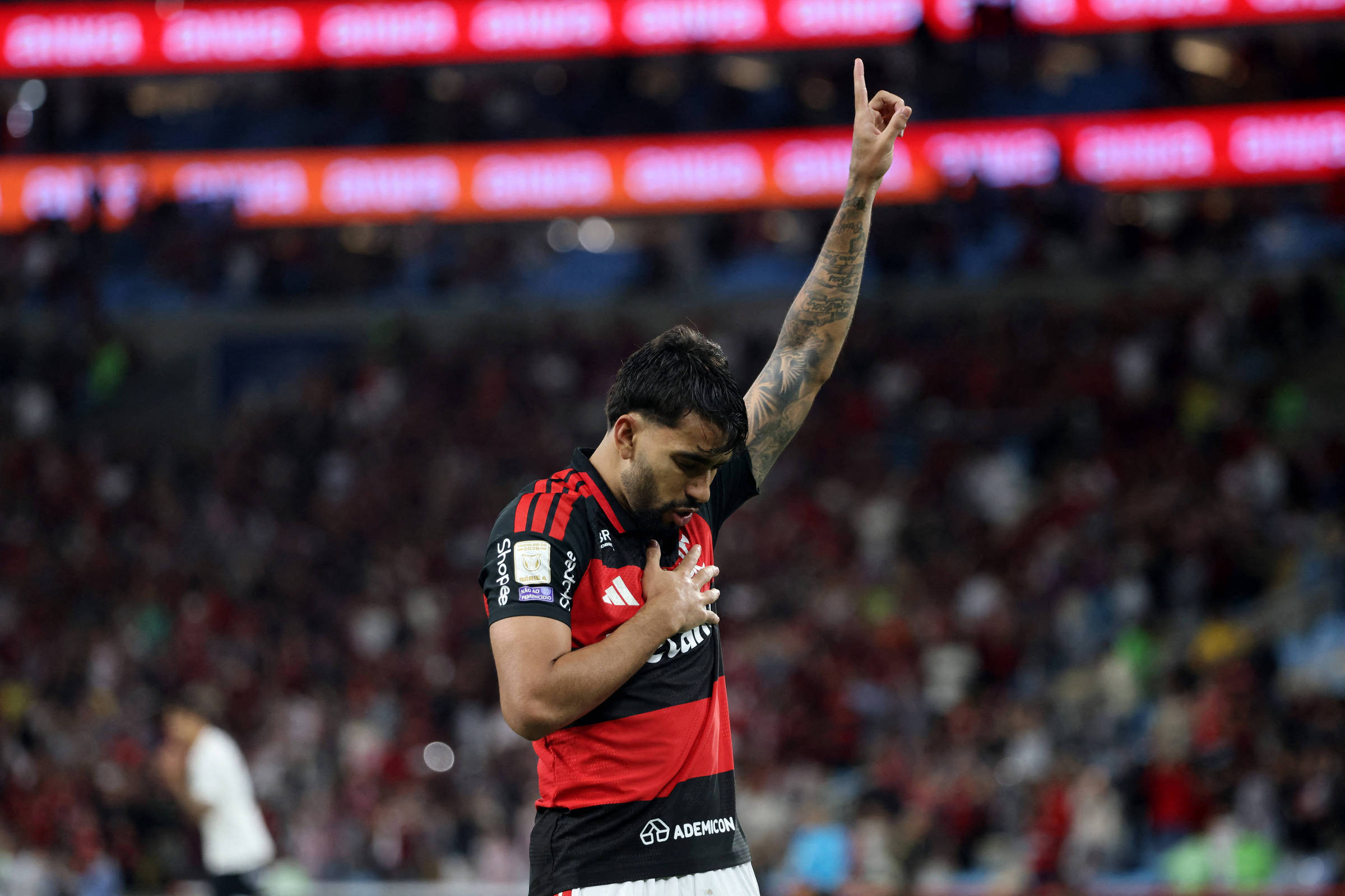 Jogador do Flamengo com uniforme vermelho e preto levanta o braço direito apontando para cima e toca o peito esquerdo com a mão esquerda, em comemoração. Estádio lotado ao fundo com torcedores desfocados.