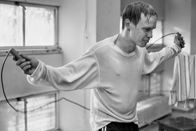 Jarkko Lahti em "O Dia Mais Feliz da Vida de Olli Mäki". A fotografia está em preto e branco. Lahti é um homem branco com cabelos loiros com alguns sinais de calvície. Ele veste uma camisa com tecido fino, de cor clara e mangas longas. Ele esta pulando corda e suando em um ginásio de esportes. 